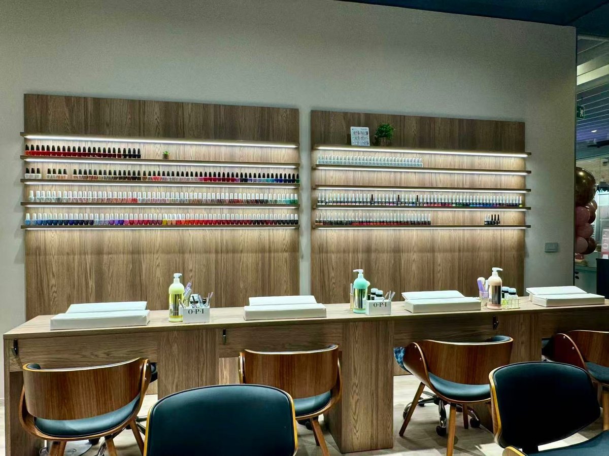 Modern salon reception area with wooden cabinetry, shelves of nail polish bottles, white counters, and upholstered chairs